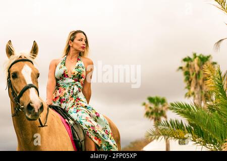 Portrait of young beautiful brunette woman wearing flowers dress riding brown horse at summer cloudy day. Stock Photo