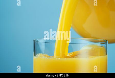 Orange juice pouring from jug into glass over blue background with copy space Stock Photo