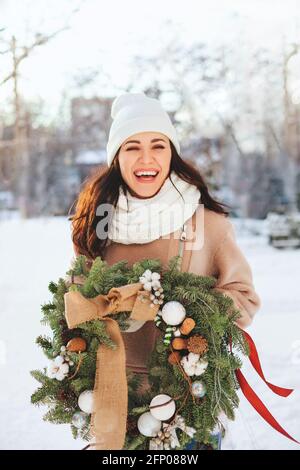 Happy female in outerwear carrying decorated Christmas wreath and standing on snow on sunny winter day Stock Photo