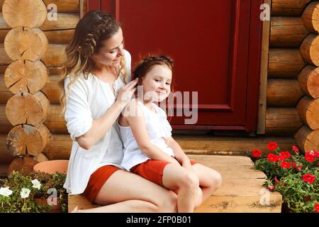 Cute little redhead girl and happy young mother with similar braided hairdo hugging together while standing against red door Stock Photo
