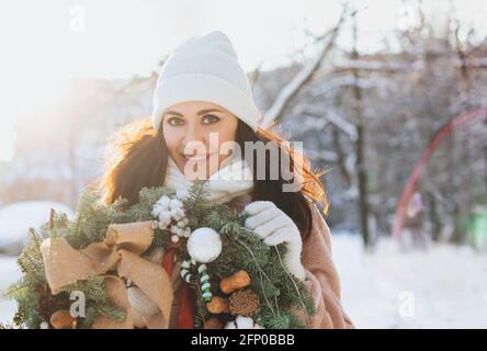 Happy female in outerwear carrying decorated Christmas wreath and standing on snow on sunny winter day Stock Photo