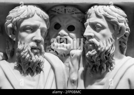 Black and white photo of ancient roman sculptures carved on a marble wall showing in detail two bearded men talking and a spooky mask between them Stock Photo