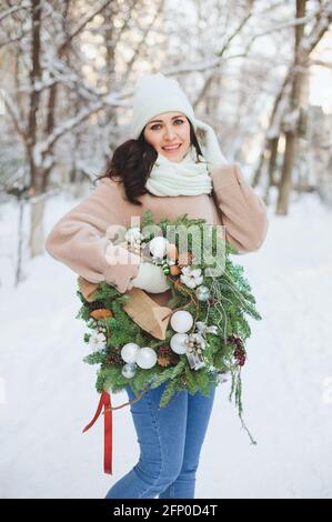 Young female in outerwear carrying decorated Christmas wreath and standing on snow on sunny winter day Stock Photo