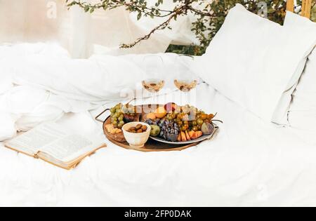 Pair of glasses with wine placed near tables with various fresh ripe fruits on blanket with cushions during romantic picnic in garden Stock Photo