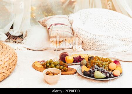 Pair of glasses with wine placed near tables with various fresh ripe fruits on blanket with cushions during romantic picnic in garden Stock Photo