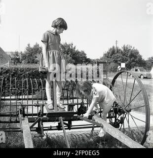 1960, historical, two young girls playing on an old agricultural, steel  made farming rake from around the early 1900s, a wire-wheeld hay rake that would have been pulled by an ox or horse, England, UK. Stock Photo