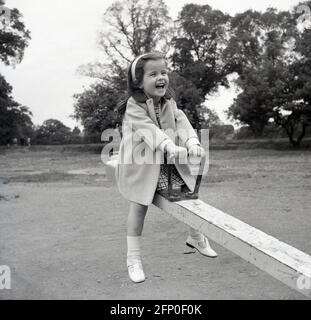 1960s, historical, outside at a park playground, a young girl having great fun riding on a metal seesaw (or see-saw) England, UK. Also known as a teeter-totter or teeterboard. A traditional piece of playground equipment, the seesaw is a long narrow metal, as seen here (or wooden) beam with a pivot point in the centre,  so as one end goes up, the other goes down, with handles eiither end to hold onto. Great fun, they were a common site in public playgrounds across Britain in this era Stock Photo