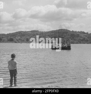 1961, historical, a young boy standing shoreside beside the calm water of a loch, watching a small cable or chain ferry taking cars and people going across the lake, Highlands of Scotland. There are hundreds of small off-shore islands in Scotland and some of the remote ones can only be reached by ferry. A cable ferry goes across a body of water pulled by cables connected to both shores, early cable ferries used rope or steel chains, hence the name of chain ferry. Stock Photo