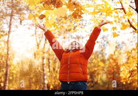 Delighted female in outerwear smiling  with dry leaves on sunny weekend day in autumn park Stock Photo