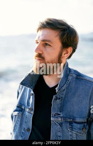 Portrait of a pensive man in a denim jacket looking into the distance against the background of the sea. Close-up Stock Photo