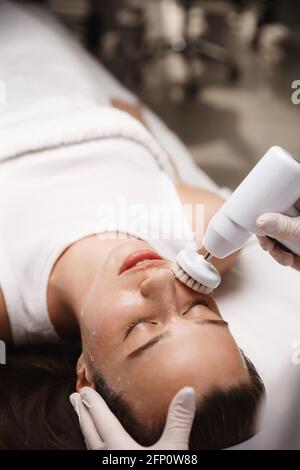 Hardware cosmetology and beauty salon. Face of young woman lying at beautician clinic, doctor using cleansing deep scrubbing rotating brush with Stock Photo