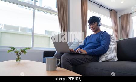 Young asian male tech user relaxing  holding laptop computer and looking at the screen in living room, Remote Job or work from home concept Stock Photo