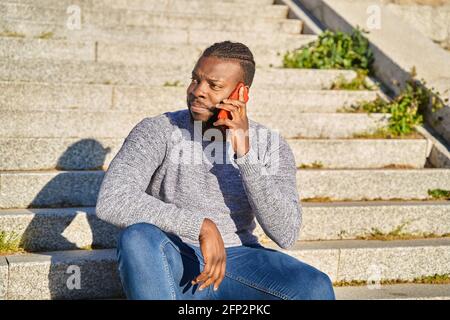 Happy black man talking on smart phone sitting on stairs. Young man in jeans and grey sweater. Concept of communication. High quality photo Stock Photo