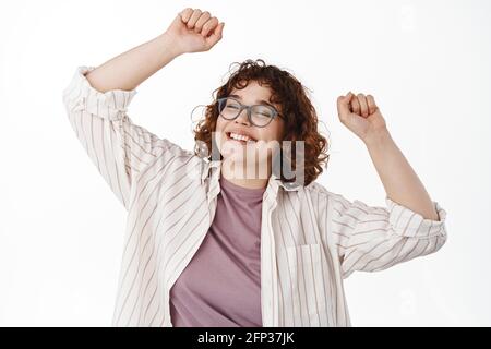 Dancing natural girl having fun, rasing hands up and partying, smiling carefree with closed eyes, relaxing in casual clothes, standing over white Stock Photo