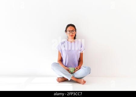 Full body of positive barefoot millennial Asian female in casual shirt and jeans sitting in light studio and drinking the coffee Stock Photo