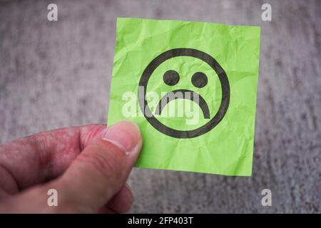 A man holding a green paper note with a sad face in his fingers. Close up. Stock Photo