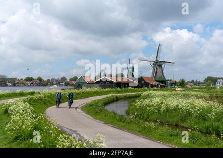 Zaandam, Netherlands - 18 May, 2021: Dutch senior citizens enjoy a bicycle ride along the canals of North Holland with traditional windmills in the ba Stock Photo