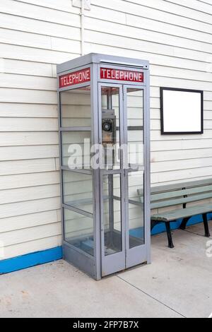 Vertical shot of an old telephone booth with a blank sign on the wall next to it on the right side.  There is a bench below the sign. Stock Photo