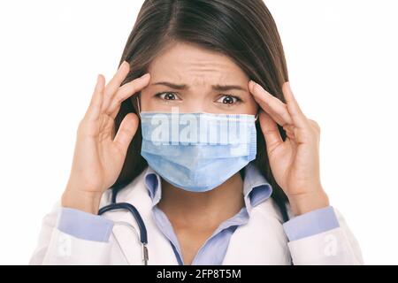 Doctor wearing coronavirus surgical face mask in panic scared doing a funny stressed out facial expression holding head from headache. Asian woman Stock Photo