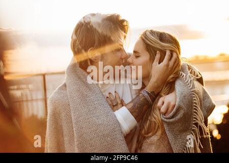 Portrait of happy beautiful couple at the sunset, cuddling in cozy blanket. Charming loving blond man hugging attractive woman, enjoying tenderness Stock Photo