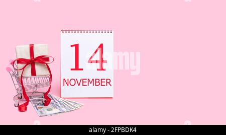 14th day of november. A gift box in a shopping trolley, dollars and a calendar with the date of 14 november on a pink background. Autumn month, day of Stock Photo