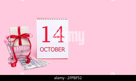 14th day of october. A gift box in a shopping trolley, dollars and a calendar with the date of 14 october on a pink background. Autumn month, day of t Stock Photo