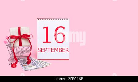 16th day of september. A gift box in a shopping trolley, dollars and a calendar with the date of 16 september on a pink background. Autumn month, day Stock Photo