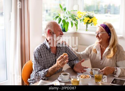 Senior couple speaking over coffee biscuits smiling touching shoulder. Man and woman in dining room relaxing. Stock Photo
