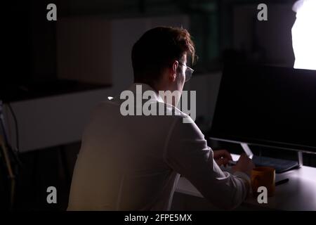 Unrecognizable Male Programmer Writing Code on Computer at Night. Side View. Young Guy In Formal Wear Engaged In Work, In Office In Dark Office. Netwo Stock Photo