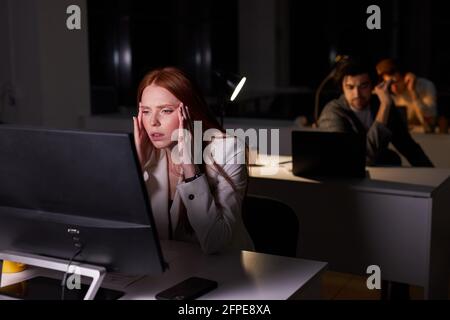 Portrait of exhausted caucasian female with red hair working in office at night, sit thinking about hard task, using pc computer, working at late with Stock Photo