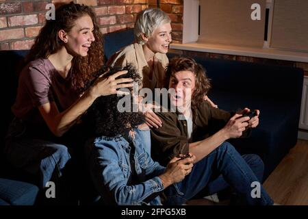 students enjoying playing videogame on game console at home holding gamepad in hands, guys on floor engaged in game while their girlfriends supporting Stock Photo