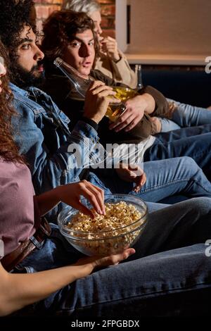 Close-up photo of friends watching comedy eating popcorn snacks in bowl and drinking beer, side view on young people watching interesting movie at hom Stock Photo