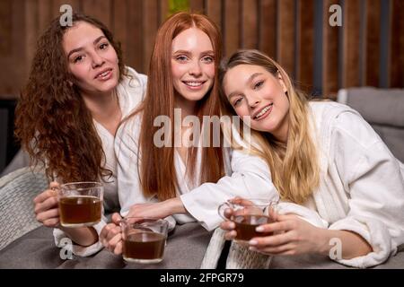 Three young ladies clothed bathrobe lying on deckchairs, talking and drinking tea, caucasian charming women having rest, leisure time, posing looking Stock Photo
