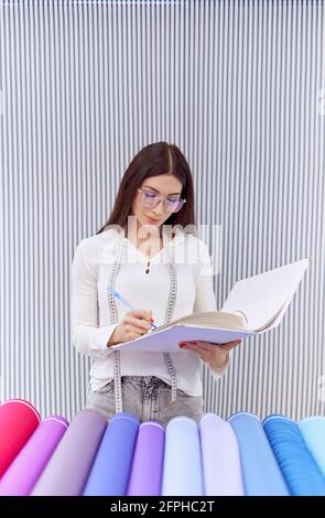 Female stylish brunette warehouse or textile store supervisor in eyeglasses making inventory in a shop. Manager calculating and making notes in journal. Concept of e-commerce or retail Stock Photo