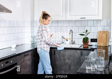 Housewife cleaning the kitchen table and sink with clot blue rag, young female in casual wear housekeeping alone at home, side view Stock Photo