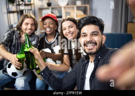 Smiling diverse people sitting together on sofa during football match and taking selfie. Concept of friendship, technology and entertainment. Stock Photo