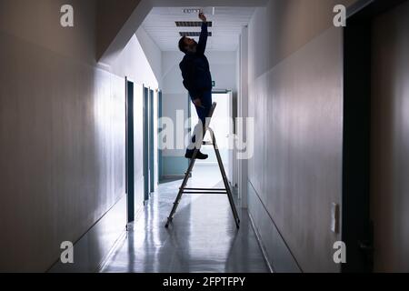 Technician Repairing Ceiling Fixture In Office On Stepladder Stock Photo