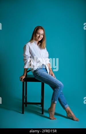 Young beautiful woman posing on a chair in a white shirt and jeans on a  blue background barefoot Stock Photo - Alamy