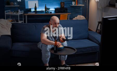 Football sport watching game late at night eating popcorn, cheering beer bottle for favourite winner team. Man enjoying championship entertainment match soccer fan looking at competition on television Stock Photo