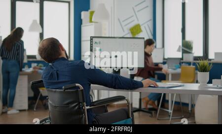Young worker with disabilities analyzing statistics datas sitting in wheelchair in front of computer talking with colleague typing at pc in startup workplace. Diverse team discussing financial project Stock Photo