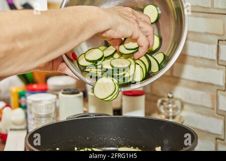 Woman puts zucchini in frying pan for making gratin with blue cheese, step-by-step recipe from the internet. Stock Photo