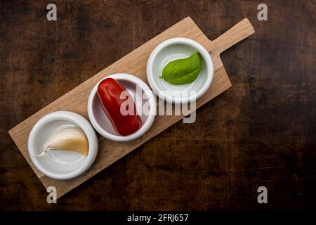 Garlic, mini tomato and a basil leaf in small white porcelain bowls on a wooden board. Stock Photo