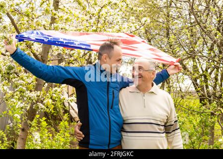 Handsome dad with his little cute sun are laying on green grassy lawn on American flag with American football ball in hand. Stock Photo