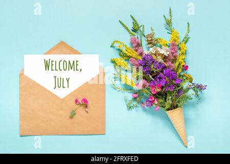 Welcome July text on paper card in craft envelope and bouquet field colored flowers in waffle ice cream cone on blue background. Creative Flat Lay Top Stock Photo