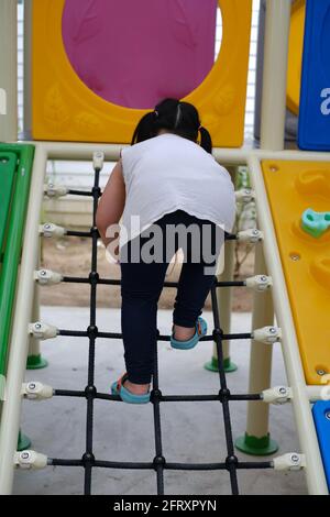 A back view of a cute young chubby Asian girl climbing up a ramp on a large colorful outdoor jungle gym, making her way to the top of the structure. Stock Photo