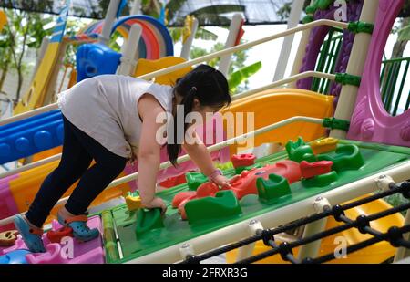 A sideview of a cute young chubby Asian girl climbing up a ramp on a large colorful outdoor jungle gym, making her way to the top of the structure. Stock Photo