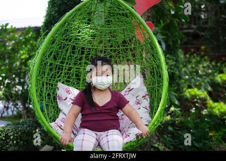 A cute young chubby Asian girl is sitting in a green round rattan swing chair with pillows in a garden, resting and relaxing. Stock Photo