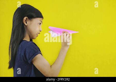 A cute young Asian girl is playing with her pink paper aero plane, holding it with one hand, aiming and getting ready to launch. Bright yellow backgro Stock Photo