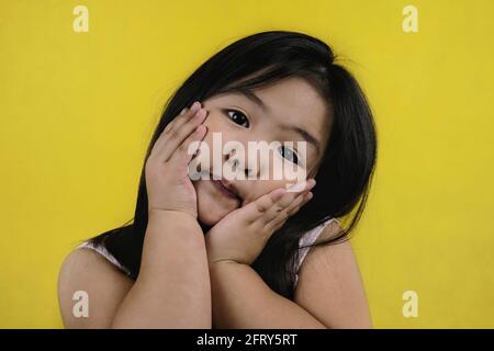 A close up picture of a cute young Asian girl holding up her face with her hands, covering her cheeks, smiling, feeling happy. Bright yellow backgroun Stock Photo