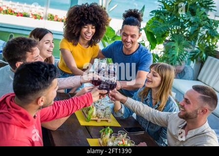 multicultural group of friends having fun and drinking wine at sunset on rooftop get together. seven young adults hanging out toasting drinks outdoors Stock Photo